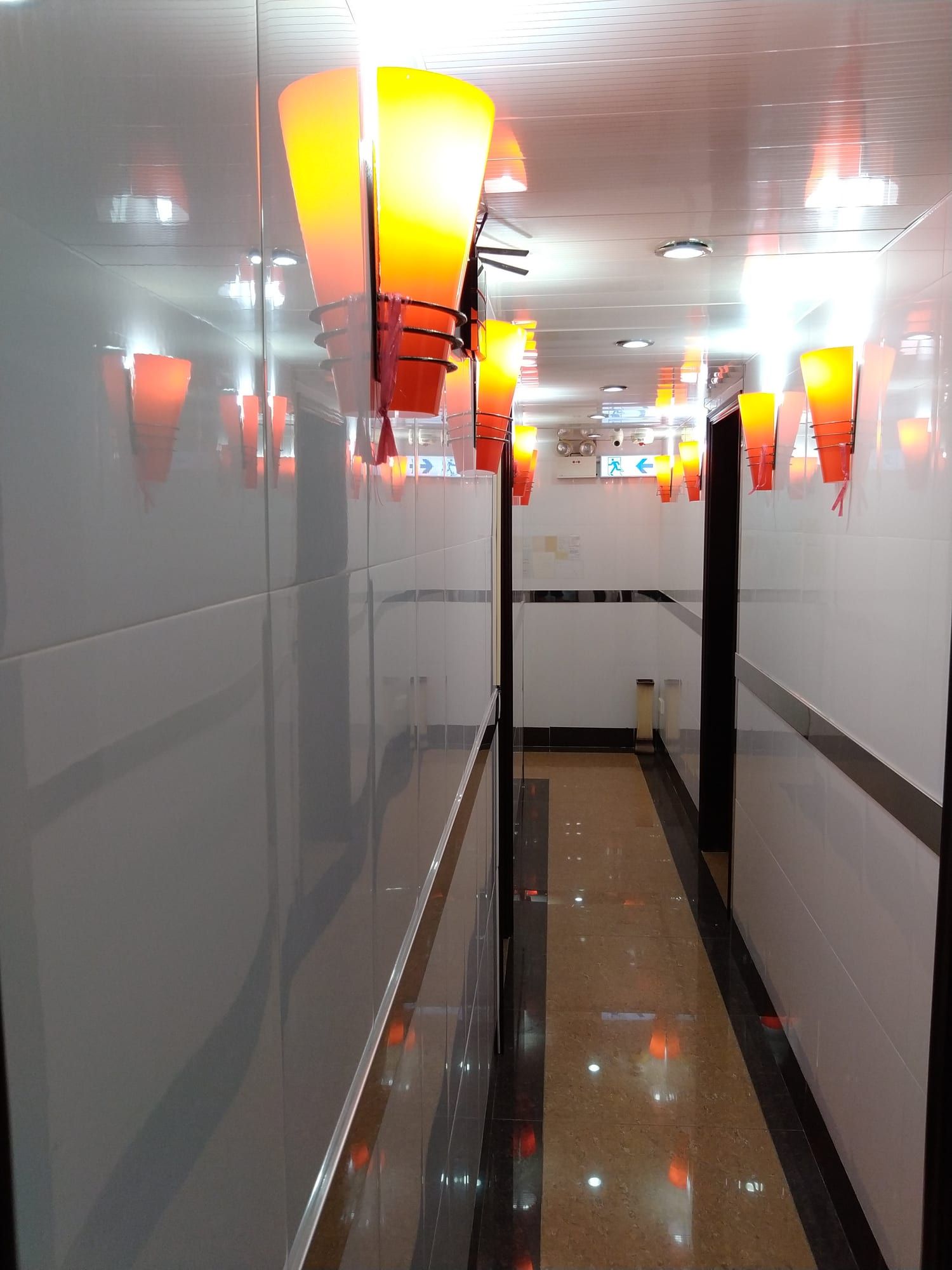 賓館走廊:白色瓷磚牆、橙色圓錐形壁燈、光亮地板,盡頭有出口指示牌。