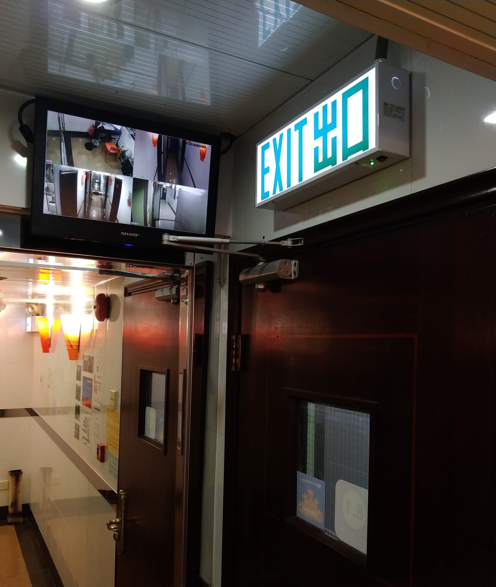 入口:深色木防火門,鐵絲網玻璃窗上貼有兩張獎項貼紙,旁有亮燈「EXIT 出口」指示牌和顯示四個鏡頭畫面的閉路電視螢幕。
